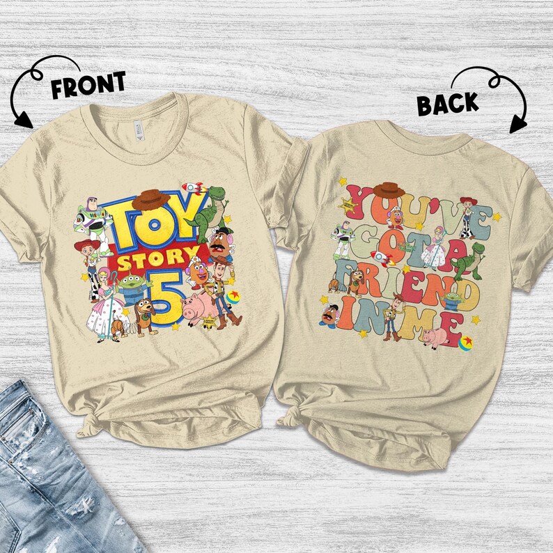 K&ouml;nnte beinhalten: Zwei beigefarbene T-Shirts mit den Aufdrucken "TOY STORY 5" und "YOU'VE GOT A FRIEND IN ME". Das vordere Shirt zeigt verschiedene Toy Story-Charaktere. Das hintere Shirt hat die gleichen Charaktere und Text in bunten Buchstaben.
