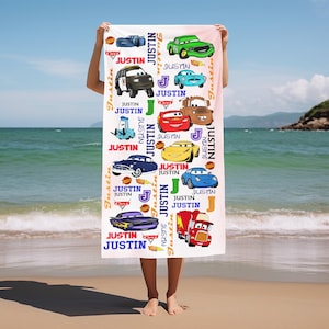 May include: A white beach towel featuring colorful cartoon cars and the name "Justin" in various fonts. The towel has a light pink background and is held up on a beach with the ocean in the background.