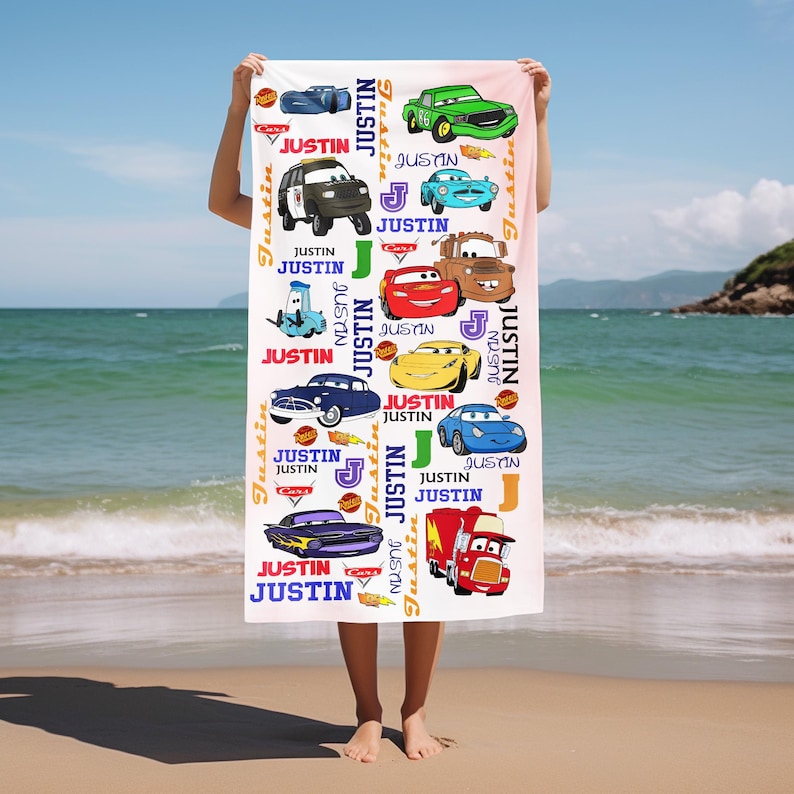 May include: A white beach towel featuring colorful cartoon cars and the name "Justin" in various fonts. The towel has a light pink background and is held up on a beach with the ocean in the background.