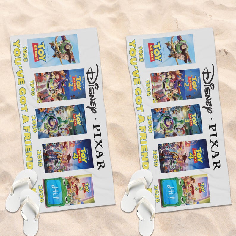 May include: A white beach towel featuring the Disney Pixar Toy Story movie franchise posters from 1995 to 2026, with the text YOU'VE GOT A FRIEND printed vertically in yellow, accompanied by a pair of white flip-flops placed at the bottom.