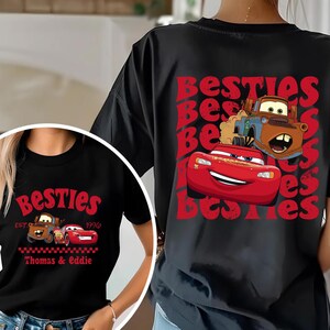 Może przedstawiać: Czarne koszulki z czerwonym nadrukiem postaci Zygzaka McQueena i Złomka z filmu Auta. Słowo "Besties" jest wydrukowane na czerwono, wraz z imionami "Thomas & Eddie".