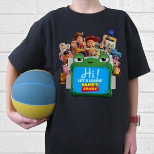 May include: Black t-shirt with a vibrant graphic of characters from the film "Toy Story." The design includes a green frog-shaped frame with the text "Hi! Let's Learn! Name's Story."