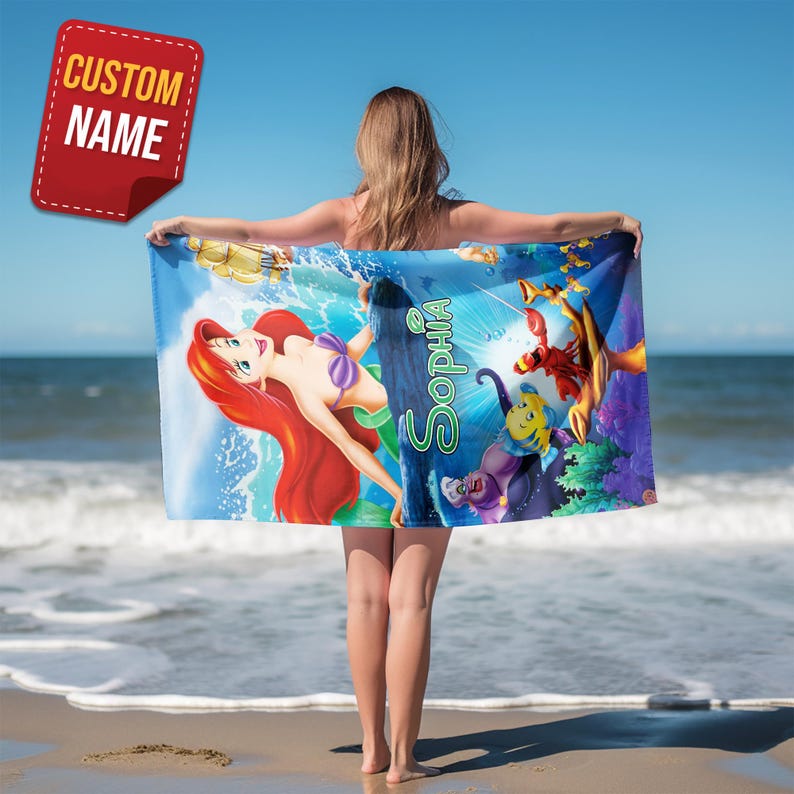 May include: A beach towel featuring a colorful illustration of Ariel from The Little Mermaid, with the name "Sophia" in green script. The towel is held up against a blue sky and ocean background, with a red "Custom Name" tag.