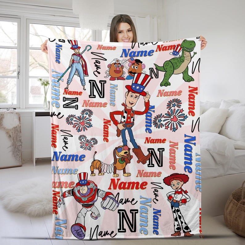 Op de afbeelding: Een lichtroze deken met stripfiguren, waaronder Woody, Buzz Lightyear en Rex. De deken heeft een herhalend patroon van het woord "Name" en de letter "N" in een scriptlettertype, met een rood-wit-blauw kleurenschema.