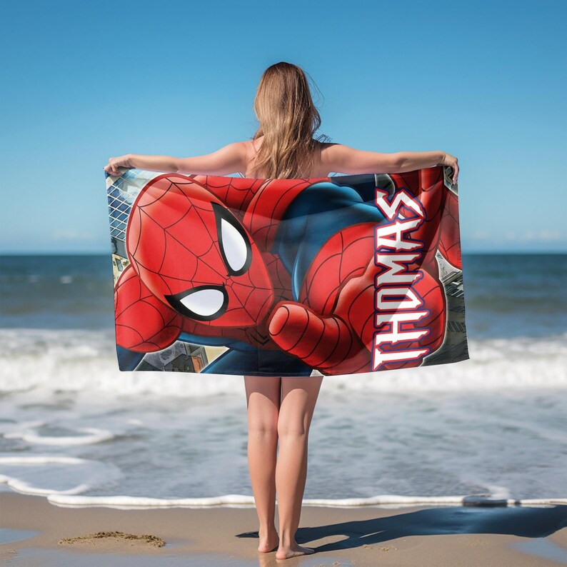 May include: A red and blue Spider-Man beach towel with the name "THOMAS" printed in red. The towel features a detailed illustration of Spider-Man and is held by a person on a sandy beach with the ocean and blue sky in the background.