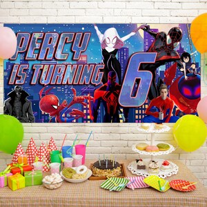 May include: A rectangular birthday banner featuring Spider-Man characters and the text Percy is turning 6, displayed above a table with party supplies including colorful balloons, gift boxes, party hats, and a chocolate cake on a stand.