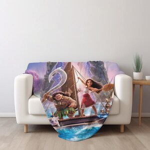 May include: A soft throw blanket featuring a colorful graphic print of Moana and Maui on a wooden sailing boat. The design includes Maui holding a large white hook, Moana holding a wooden paddle, and a small pig character on the deck.