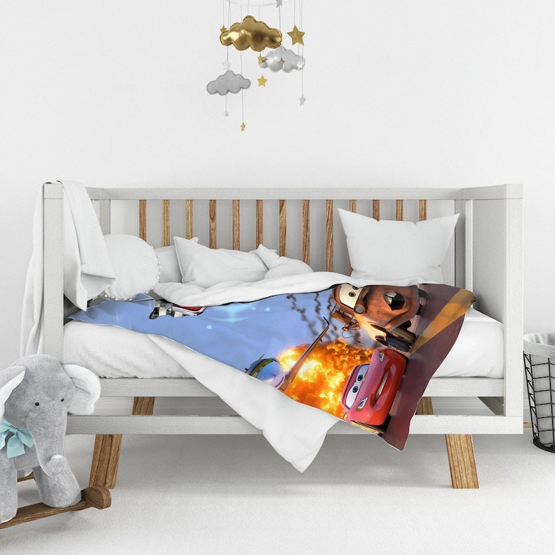 May include: A soft throw blanket featuring a vibrant graphic print of characters from the movie Cars, including a red race car and a brown tow truck, set against a blue sky background with a large orange explosion and scattered debris.