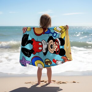May include: A light blue beach towel featuring Mickey Mouse wearing red sunglasses and shorts, surrounded by beach-themed illustrations. The towel is held by a person on a sandy beach with the ocean in the background.