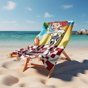 May include: A beach towel draped over a wooden lounge chair featuring a cartoon character with red hair, a white shirt, and a cow-print vest. The towel has a red and white checkered pattern at the bottom with the name Jessie printed in a blue border.