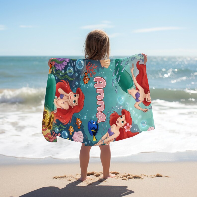 Peut inclure: Une serviette de plage avec Ariel, la Petite Sir&egrave;ne, aux cheveux roux et un soutien-gorge en coquillage violet, nageant dans une sc&egrave;ne sous-marine avec des poissons et des bulles. Le nom "Anna" est imprim&eacute; en rose. La serviette est tenue par une personne sur une plage de sable.