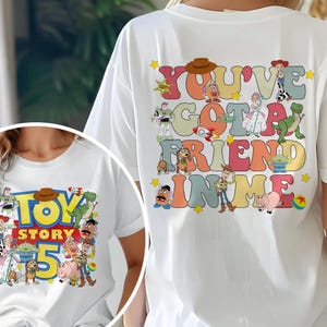 K&ouml;nnte beinhalten: Wei&szlig;es T-Shirt mit einer farbenfrohen Grafik von Figuren und Text aus dem Film "Toy Story". Der Text auf der R&uuml;ckseite des Shirts lautet "You've Got a Friend in Me". Auf der Vorderseite des Shirts steht "Toy Story 5".