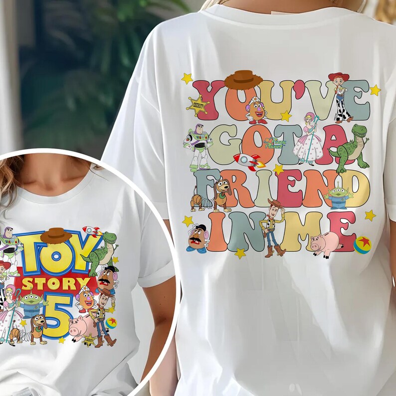 K&ouml;nnte beinhalten: Wei&szlig;es T-Shirt mit einer farbenfrohen Grafik von Figuren und Text aus dem Film "Toy Story". Der Text auf der R&uuml;ckseite des Shirts lautet "You've Got a Friend in Me". Auf der Vorderseite des Shirts steht "Toy Story 5".