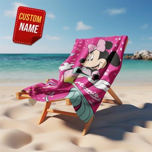 Pode incluir: Uma toalha de praia rosa com Minnie Mouse como sereia, drapeada sobre uma cadeira de praia de madeira em uma praia de areia. A toalha tem um padr&atilde;o de estrelas repetidas e as palavras "Minnie mermaid". Uma etiqueta vermelha com as palavras "CUSTOM NAME" est&aacute; no canto superior esquerdo.