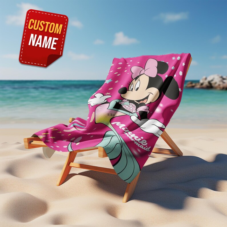 Pode incluir: Uma toalha de praia rosa com Minnie Mouse como sereia, drapeada sobre uma cadeira de praia de madeira em uma praia de areia. A toalha tem um padr&atilde;o de estrelas repetidas e as palavras "Minnie mermaid". Uma etiqueta vermelha com as palavras "CUSTOM NAME" est&aacute; no canto superior esquerdo.