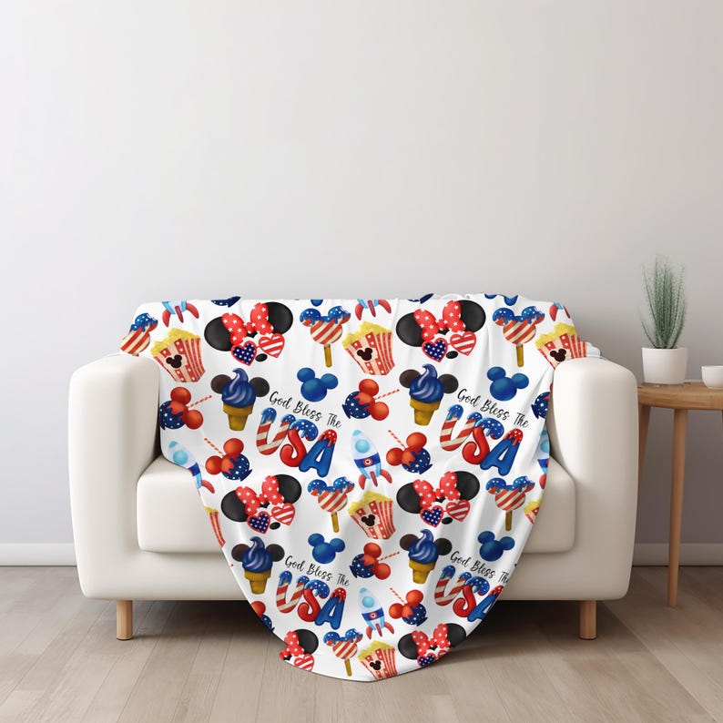 Puede incluir: Una manta blanca con un patr&oacute;n repetido de ilustraciones patri&oacute;ticas. El dise&ntilde;o incluye orejas de Mickey Mouse, conos de helado, palomitas de ma&iacute;z, cohetes y el texto "God Bless The USA" en rojo, blanco y azul.