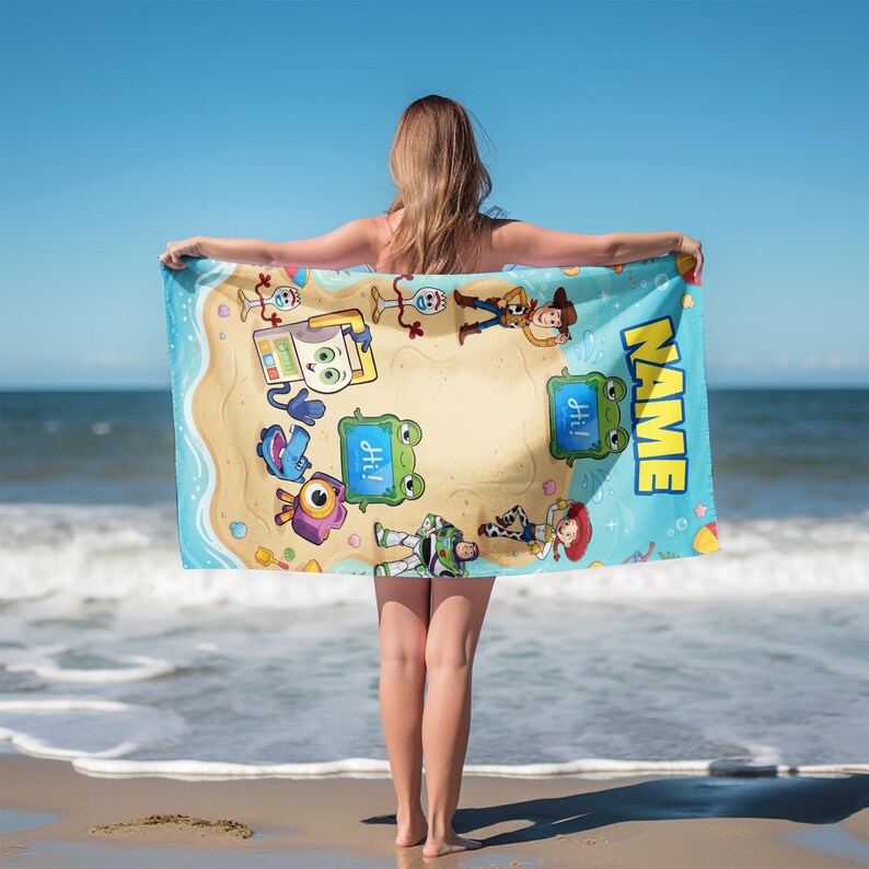 May include: A rectangular beach towel featuring cartoon characters on a sandy island design with a blue border. The towel has the word "NAME" printed on it. The characters include Woody, Buzz Lightyear, and others. The word "Hi!" is also printed on the towel.