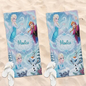 K&ouml;nnte beinhalten: Zwei Strandt&uuml;cher mit Elsa, Anna und Olaf aus Frozen, mit dem Namen "Monica". Die T&uuml;cher haben ein hellblaues und lila Winterlandschaftsdesign. Wei&szlig;e Flip-Flops liegen auf einer sandigen Oberfl&auml;che neben den T&uuml;chern.