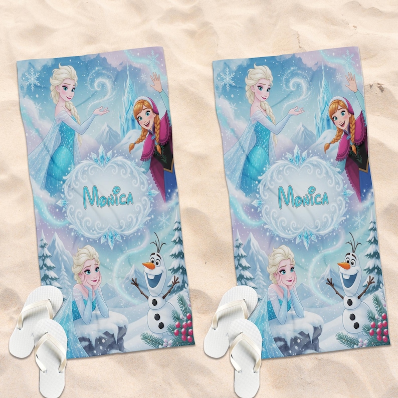 K&ouml;nnte beinhalten: Zwei Strandt&uuml;cher mit Elsa, Anna und Olaf aus Frozen, mit dem Namen "Monica". Die T&uuml;cher haben ein hellblaues und lila Winterlandschaftsdesign. Wei&szlig;e Flip-Flops liegen auf einer sandigen Oberfl&auml;che neben den T&uuml;chern.