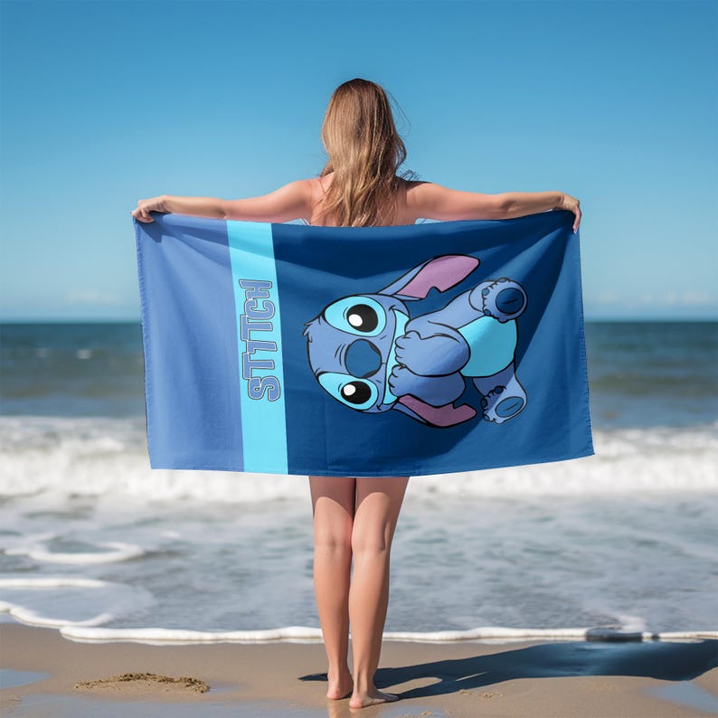 May include: A rectangular beach towel featuring a dark blue background with a light blue vertical stripe on the left side. The word STITCH is printed vertically in light blue letters. A cartoon character of Stitch is centered on the towel.