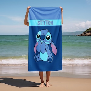 May include: A rectangular beach towel featuring a blue gradient background and the character Stitch centered in the lower portion. The word STITCH is printed in a light blue font across a horizontal band near the top of the towel.