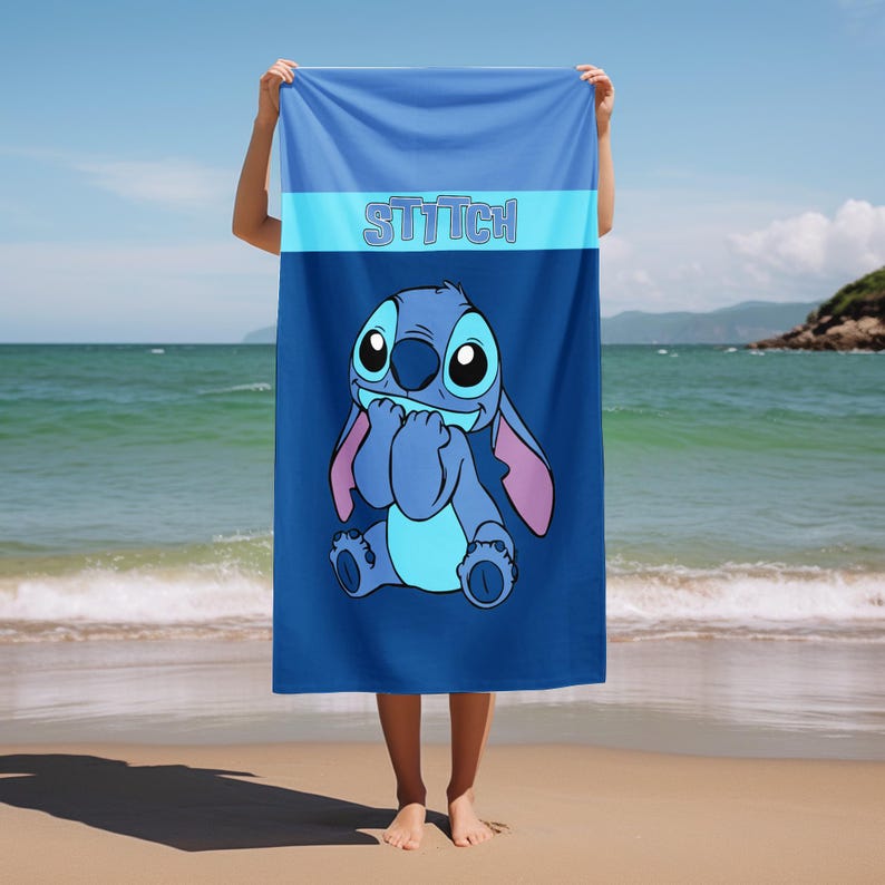 May include: A rectangular beach towel featuring a blue gradient background and the character Stitch centered in the lower portion. The word STITCH is printed in a light blue font across a horizontal band near the top of the towel.