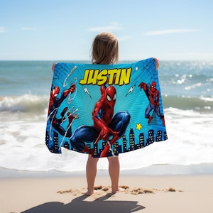 Op de afbeelding: Blauw strandlaken met Spider-Man illustraties en de naam "JUSTIN" in geel. De handdoek heeft een silhouet van een stadsgezicht aan de onderkant en een ster. De achtergrond is helderblauw met webontwerpen.
