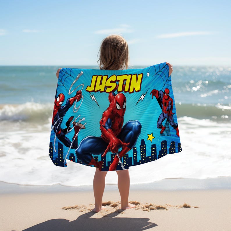 Op de afbeelding: Blauw strandlaken met Spider-Man illustraties en de naam "JUSTIN" in geel. De handdoek heeft een silhouet van een stadsgezicht aan de onderkant en een ster. De achtergrond is helderblauw met webontwerpen.