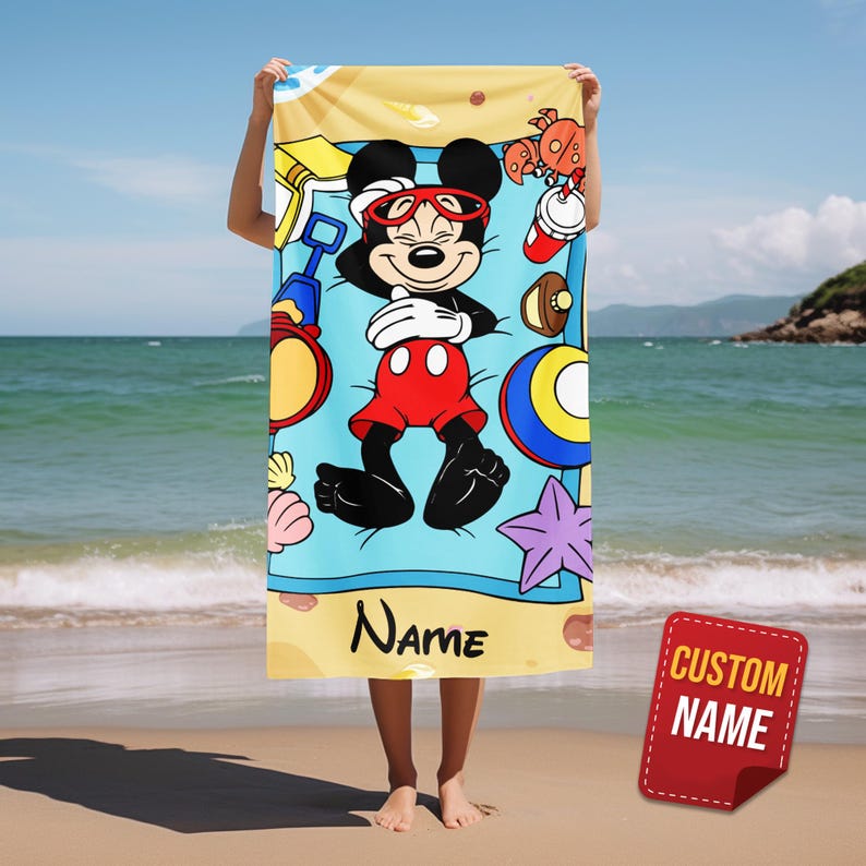 May include: A beach towel featuring Mickey Mouse in a beach scene. Mickey is wearing sunglasses and red shorts, surrounded by beach toys and a starfish. The towel has a blue background with the word "Name" printed on it.