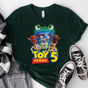 May include: Dark green t-shirt featuring a colorful graphic of characters from the movie Toy Story 5. The graphic includes Woody, Buzz Lightyear, and Jessie. The text "TOY STORY 5" is in yellow and red. Black canvas sneakers are next to the shirt.