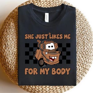 Puede incluir: Una camiseta negra de cuello redondo con un gr&aacute;fico de una gr&uacute;a de dibujos animados marr&oacute;n con auriculares. El texto "SHE JUST LIKES ME FOR MY BODY" est&aacute; impreso en marr&oacute;n sobre y debajo del cami&oacute;n, sobre un fondo de tablero de ajedrez negro y gris.