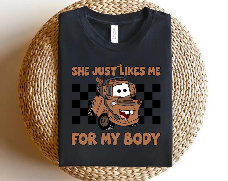 Puede incluir: Una camiseta negra de cuello redondo con un gr&aacute;fico de una gr&uacute;a de dibujos animados marr&oacute;n con auriculares. El texto "SHE JUST LIKES ME FOR MY BODY" est&aacute; impreso en marr&oacute;n sobre y debajo del cami&oacute;n, sobre un fondo de tablero de ajedrez negro y gris.