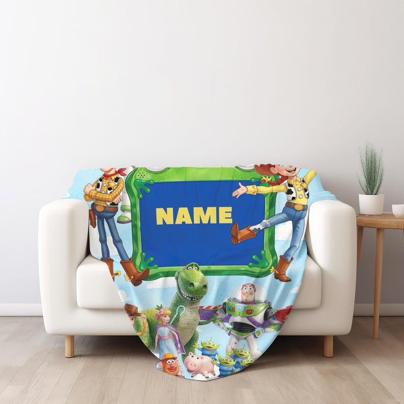 May include: A light blue fleece blanket featuring a central green rectangular frame with the word NAME in yellow text. The design includes various animated characters such as a cowboy, a cowgirl, a green dinosaur, a space ranger, and a pig.