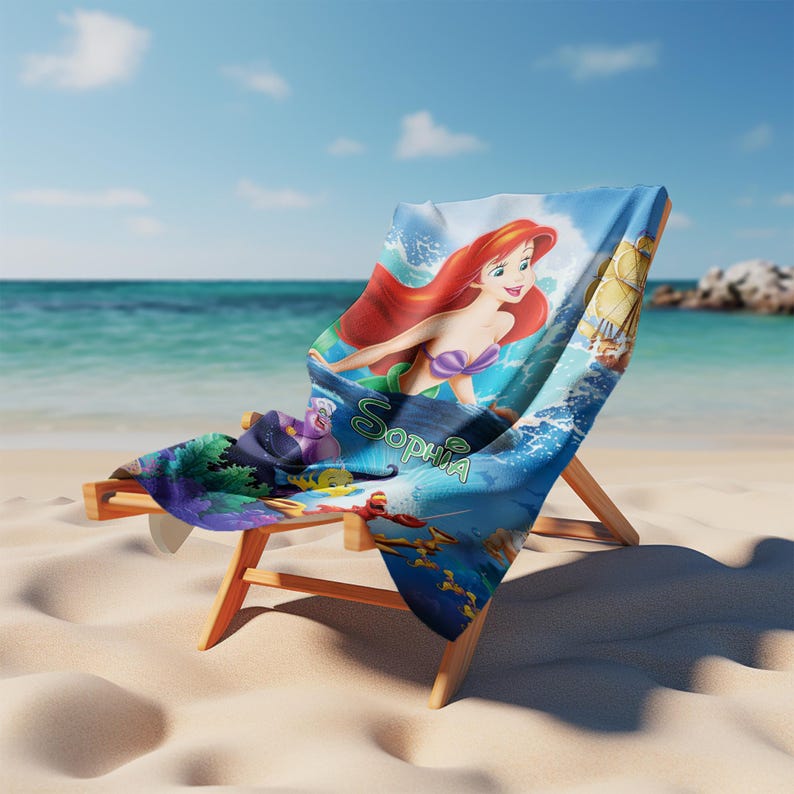 May include: A wooden beach chair with a blue and white Little Mermaid-themed towel draped over it. The towel features Ariel, the mermaid, and the name "Sophia" in green script. The chair is on a sandy beach with the ocean in the background.