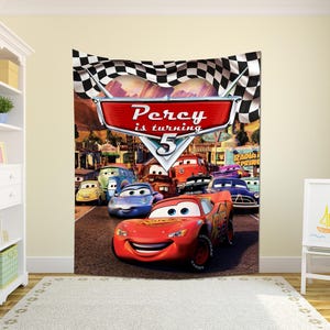 May include: A rectangular wall tapestry featuring a checkered flag border and a central red sign with the text Percy is turning 5. The scene depicts various animated car characters on a road, including a red race car in the foreground.