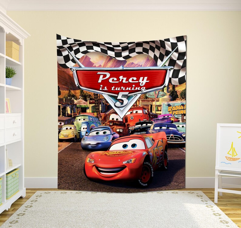 May include: A rectangular wall tapestry featuring a checkered flag border and a central red sign with the text Percy is turning 5. The scene depicts various animated car characters on a road, including a red race car in the foreground.