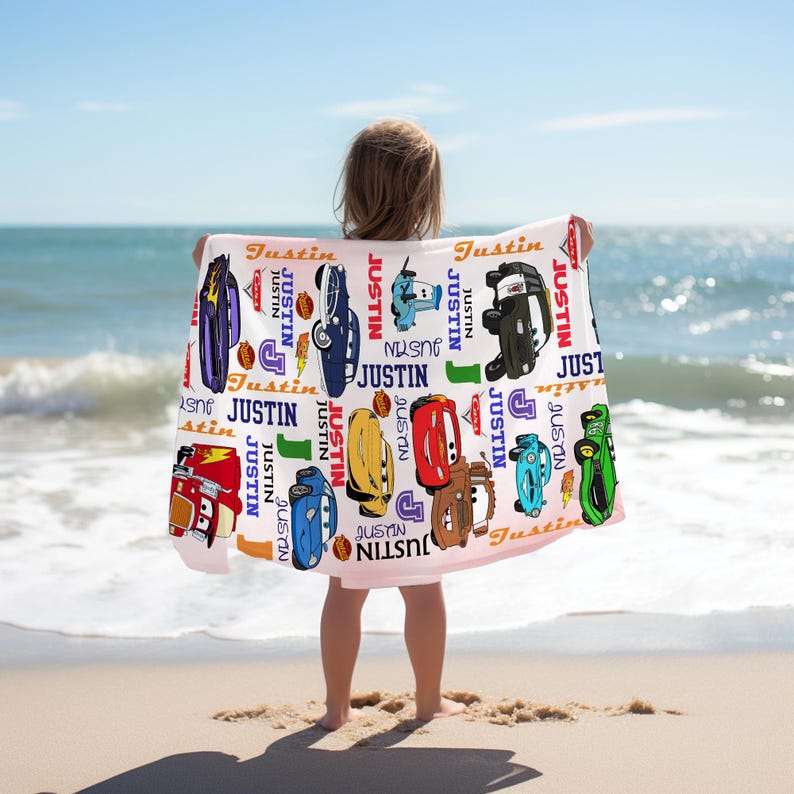May include: A personalized beach towel held by a child on a sandy beach. The towel is light pink and features various cartoon car illustrations and the name "Justin" in multiple colors and fonts.