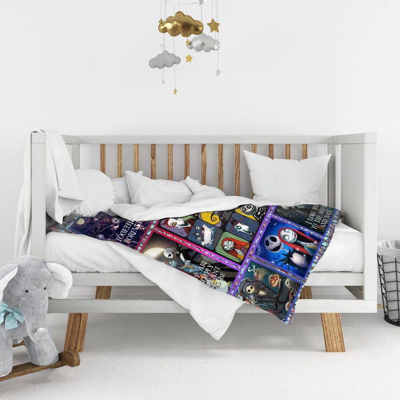 May include: A colorful throw blanket featuring a patchwork design with images of characters from The Nightmare Before Christmas. The blanket is draped over a white crib with a plush elephant toy and a mobile hanging above.