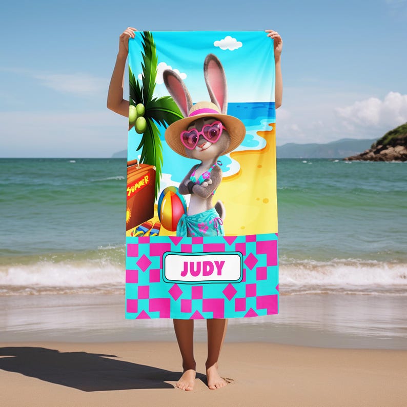 Może przedstawiać: Prostokątny ręcznik plażowy z rysunkowym kr&oacute;likiem w słomkowym kapeluszu, okularach w kształcie serc, g&oacute;rze od bikini i pareo. Tło przedstawia plażę, palmę i piłkę. W dolnej części znajduje się r&oacute;żowo-niebieski wz&oacute;r z napisem JUDY.