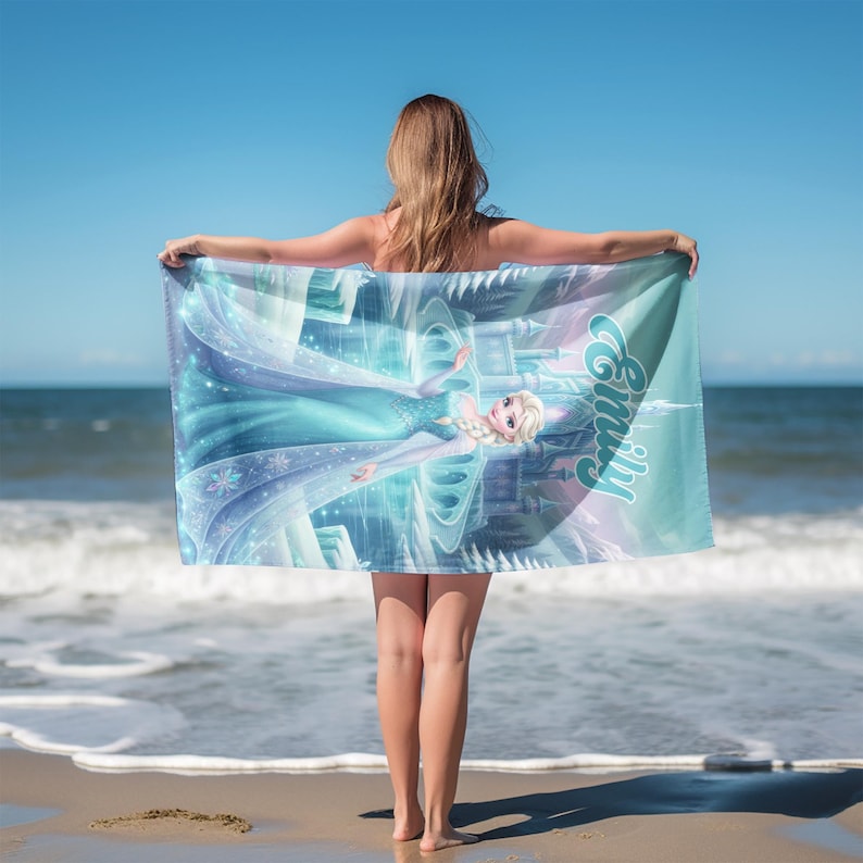 May include: A rectangular beach towel featuring a printed illustration of a blonde character in a light blue gown with a flowing cape, set against an icy castle background. The name Emily is printed in a stylised script font on the right side.