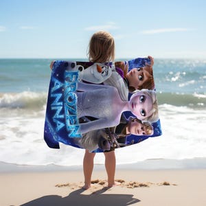 May include: A blue beach towel featuring characters from the animated film "Frozen", including Elsa, Anna, Olaf, and Kristoff. The towel has the words "Frozen" and "Anna" in a blue, sparkling font. The background is a starry night.