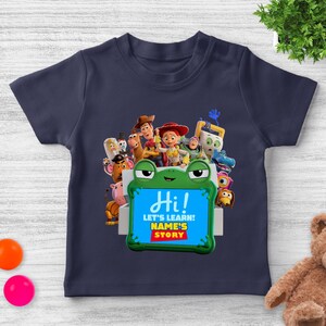 May include: Navy blue t-shirt featuring characters from the animated movie Toy Story. The shirt has a green frog-shaped frame with the word "Hi!" and the text "LET'S LEARN! NAME'S STORY" printed on it. The shirt is designed for children.