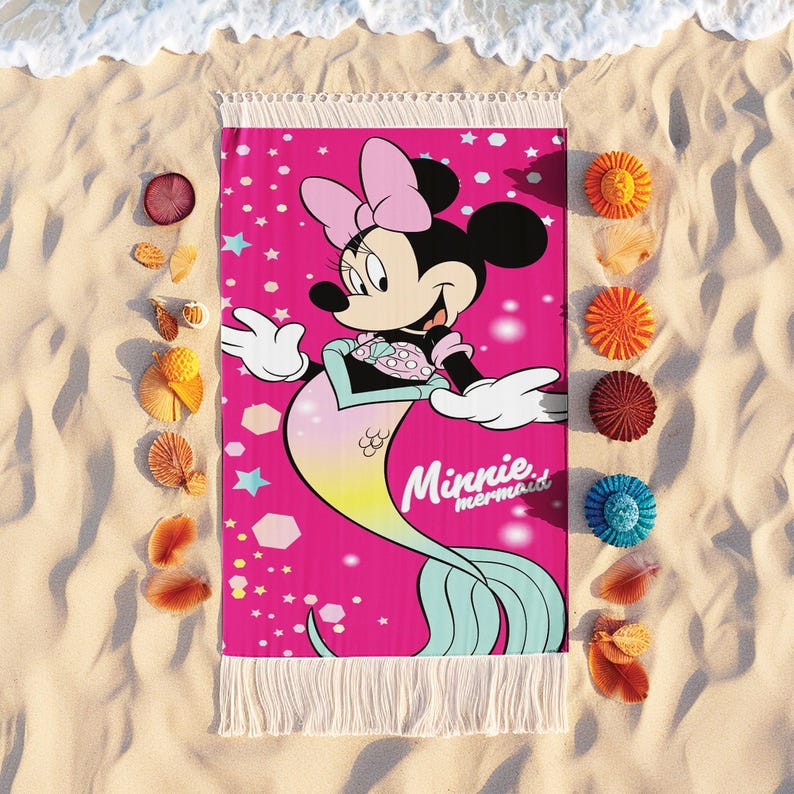 Pode incluir: Uma toalha de praia rosa com Minnie Mouse como sereia, com um la&ccedil;o rosa e uma cauda turquesa. A toalha tem franjas brancas e as palavras "Minnie mermaid". A toalha &eacute; rodeada por conchas e objetos decorativos numa praia arenosa.