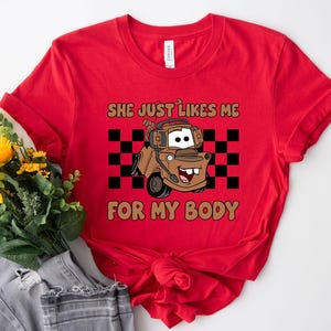 Puede incluir: Una camiseta roja de manga corta con el gr&aacute;fico de una gr&uacute;a de remolque marr&oacute;n con auriculares. El dise&ntilde;o incluye un fondo de cuadros negros y blancos y el texto "SHE JUST LIKES ME FOR MY BODY" en letras de color marr&oacute;n.