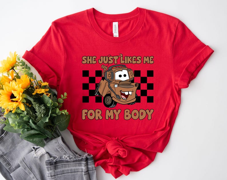 Puede incluir: Una camiseta roja de manga corta con el gr&aacute;fico de una gr&uacute;a de remolque marr&oacute;n con auriculares. El dise&ntilde;o incluye un fondo de cuadros negros y blancos y el texto "SHE JUST LIKES ME FOR MY BODY" en letras de color marr&oacute;n.