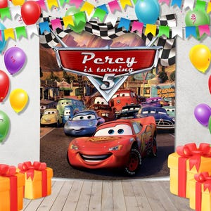 May include: A rectangular party backdrop featuring a Cars movie theme with a central red sign reading Percy is turning 5, surrounded by colorful balloons, pennant banners, and yellow gift boxes with red ribbons on a wooden floor surface.