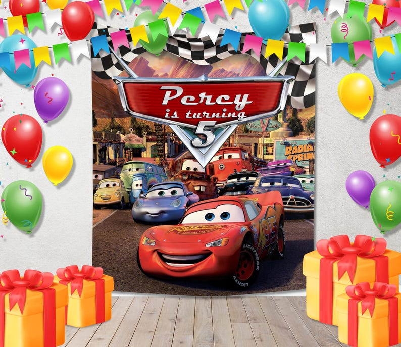 May include: A rectangular party backdrop featuring a Cars movie theme with a central red sign reading Percy is turning 5, surrounded by colorful balloons, pennant banners, and yellow gift boxes with red ribbons on a wooden floor surface.