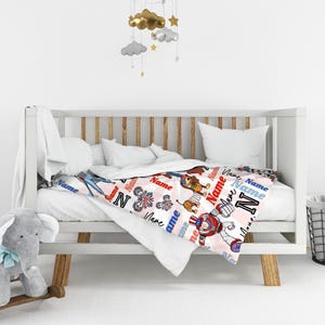 Op de afbeelding: Een babydeken met een roze achtergrond en een herhalend patroon van het woord "Name" in rood, wit en blauw. De deken heeft cartoon illustraties van een hond, een robot en andere ontwerpen. De deken is over een witte wieg gedrapeerd.