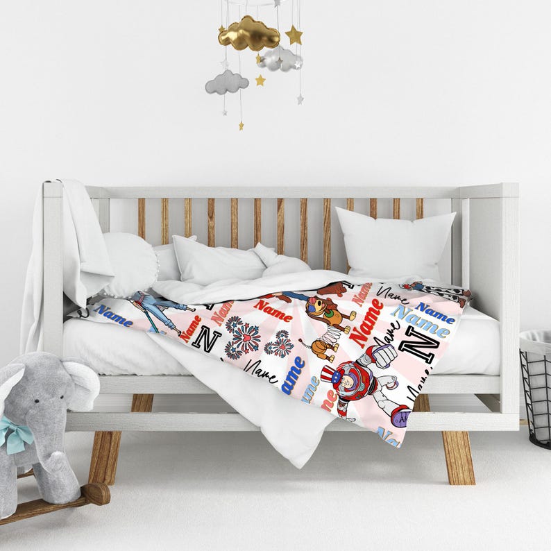 Op de afbeelding: Een babydeken met een roze achtergrond en een herhalend patroon van het woord "Name" in rood, wit en blauw. De deken heeft cartoon illustraties van een hond, een robot en andere ontwerpen. De deken is over een witte wieg gedrapeerd.