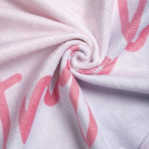May include: A soft, folded pink and white towel with a swirling pattern. The towel features large, stylized pink lettering and a striped design. The overall color scheme is a light pink and white, creating a gentle and inviting aesthetic.